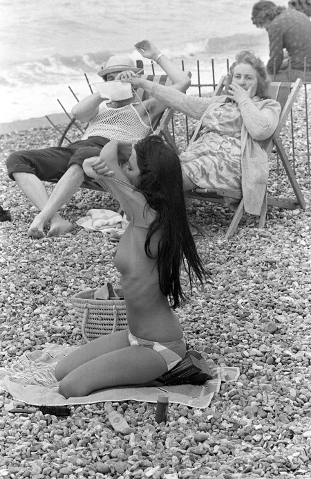 Woman covering man's eyes with her knitting at sight of young woman taking off her top on the beach in France, 1974 by maxwe11d